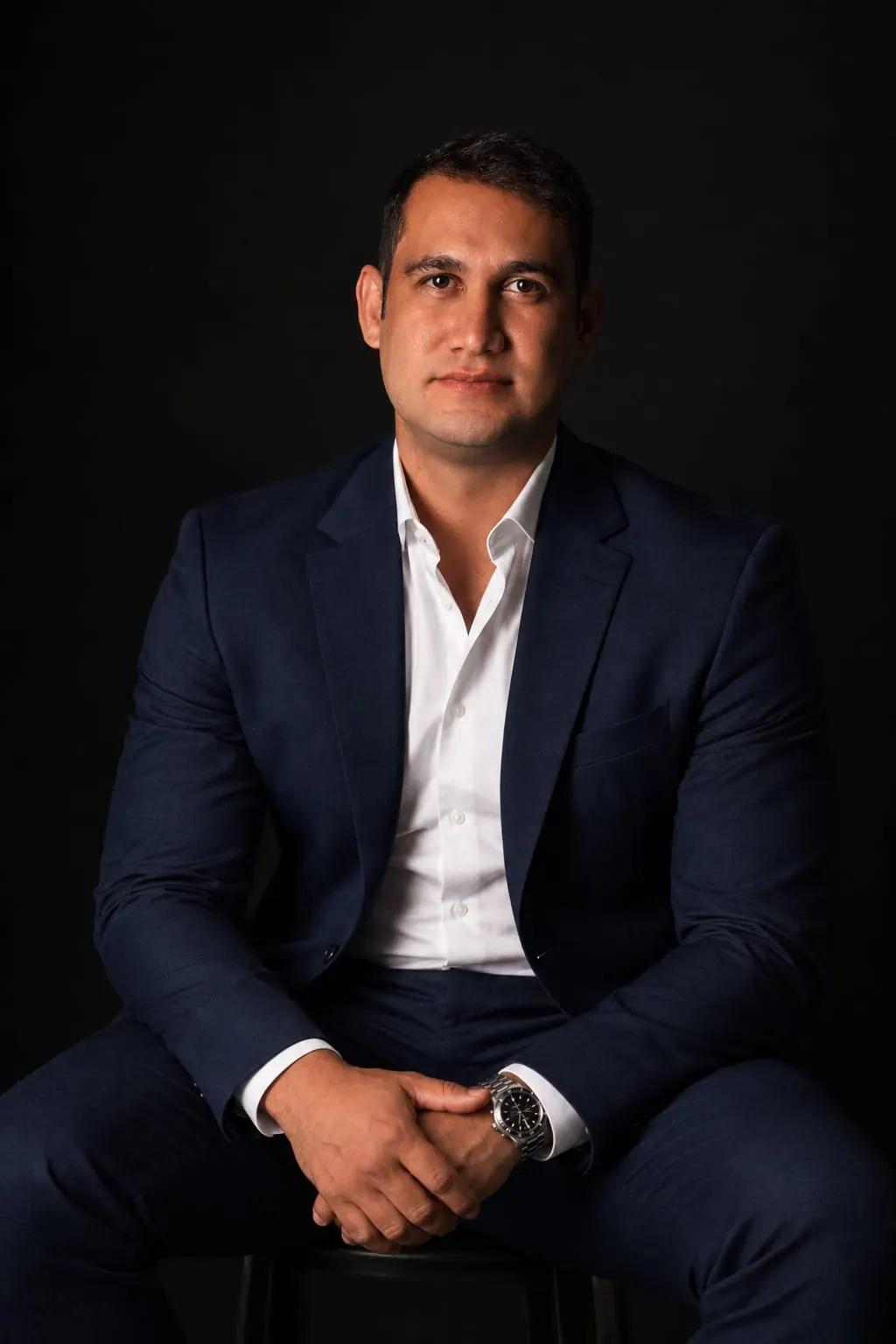 Hermeson Pires sentado, de terno azul-marinho e camisa branca, em fundo escuro